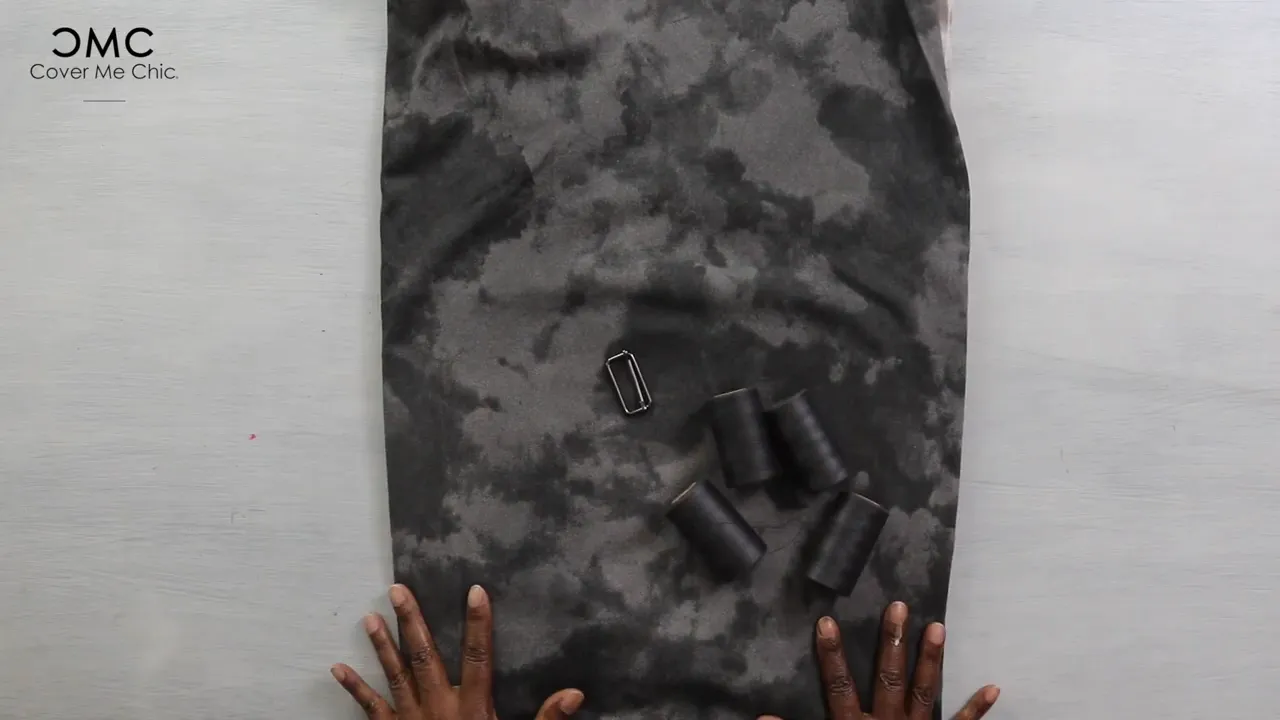 Top-down view of patterned interlock fabric with multiple black thread spools and a metal belt slider, hands framing the bottom of the shot.