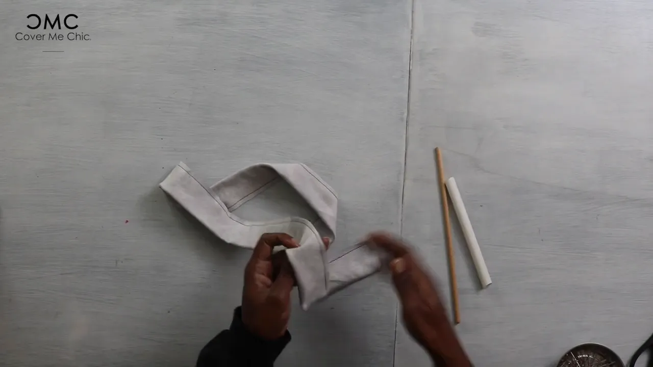 Hands holding a folded narrow fabric belt on a work surface with a wooden dowel and a white loop-turner tube nearby.